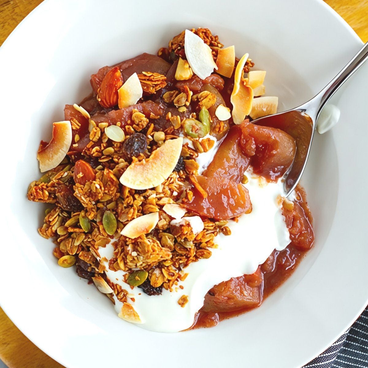 Tender apples stewed in warm mulled wine served over creamy yogurt and topped with crunchy granola, seed clusters, and toasted coconut flakes in a white bowl.