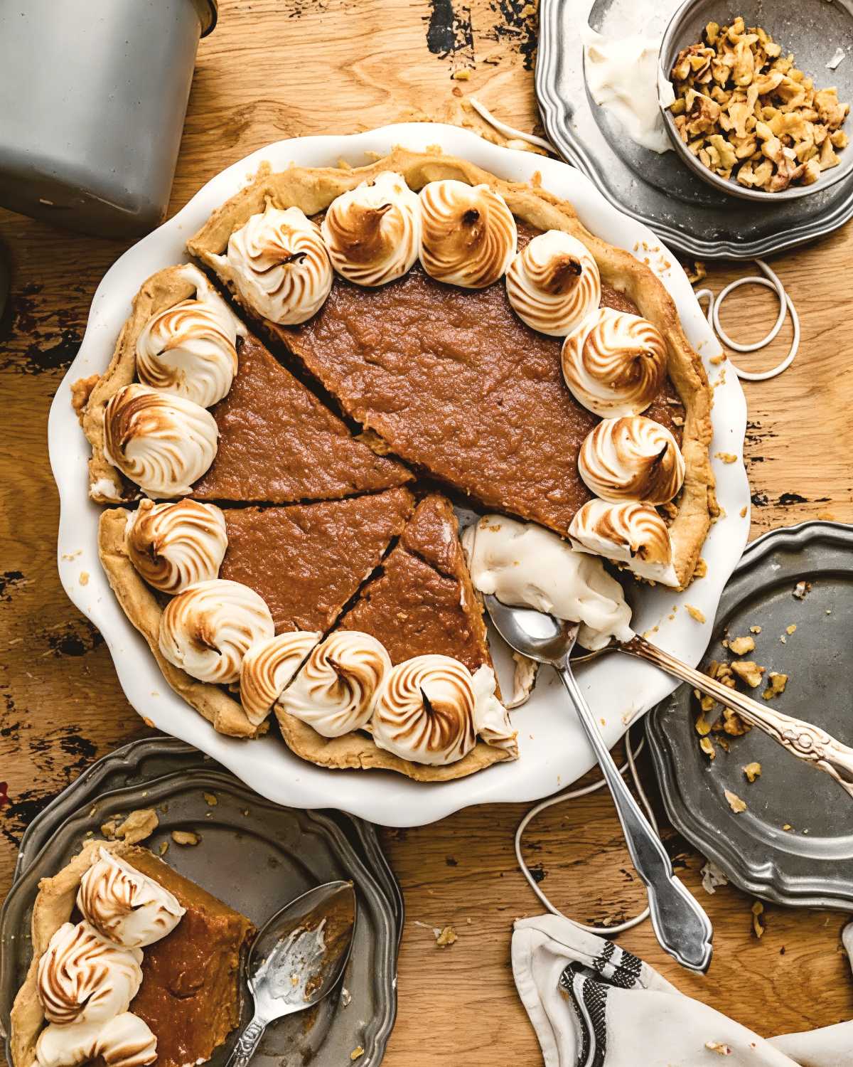An old-fashioned sweet potato pie sliced in a pie dish, elegantly bordered with toasted meringue dollops and served on a rustic wooden table alongside chopped walnuts.