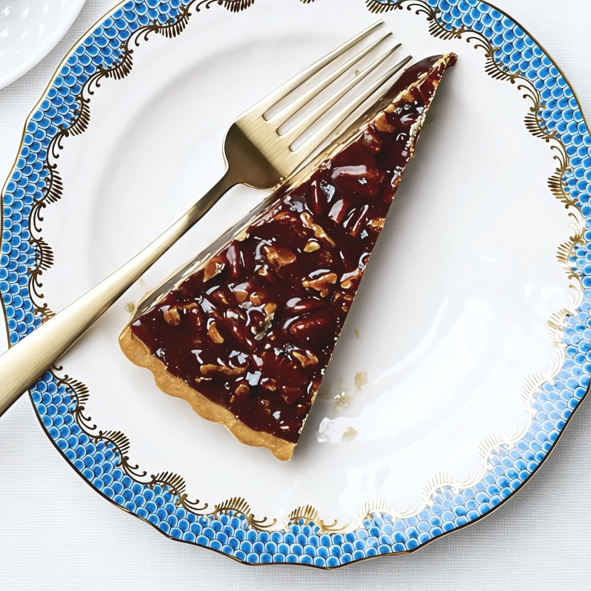 Slice of decadent chocolate caramel pecan tart served on an ornate blue and white plate with a gold fork, ideal for Thanksgiving dessert ideas.