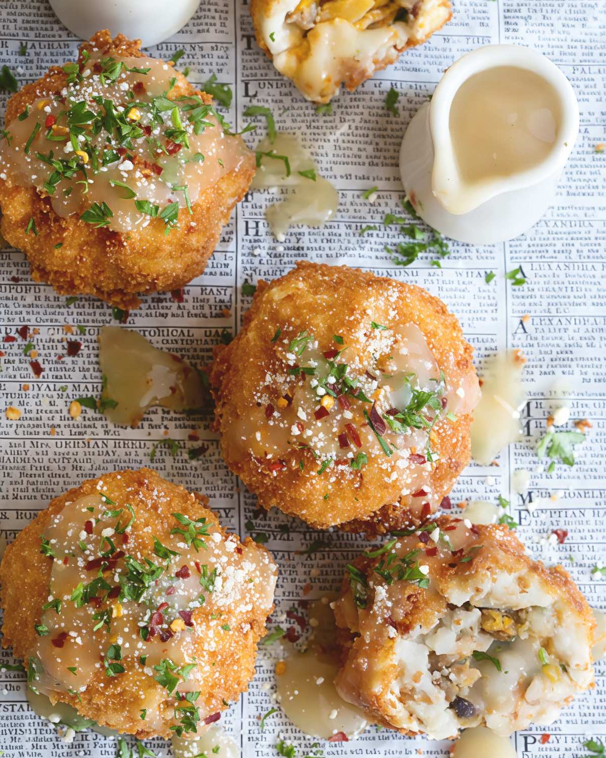 Crispy fried Thanksgiving leftover balls drizzled with savory gravy and sprinkled with fresh herbs served alongside small gravy boats on newspaper wrapping