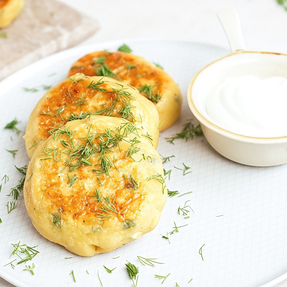 Crispy golden-brown mashed potato patties garnished with fresh dill on a plate alongside a small ramekin of sour cream.
