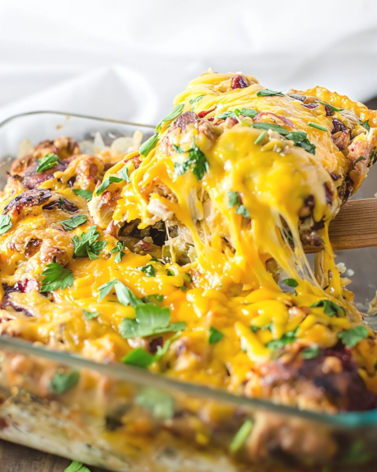 A wooden spatula lifting a serving of cheesy Thanksgiving leftover savory bread pudding topped with fresh parsley from a glass baking dish.