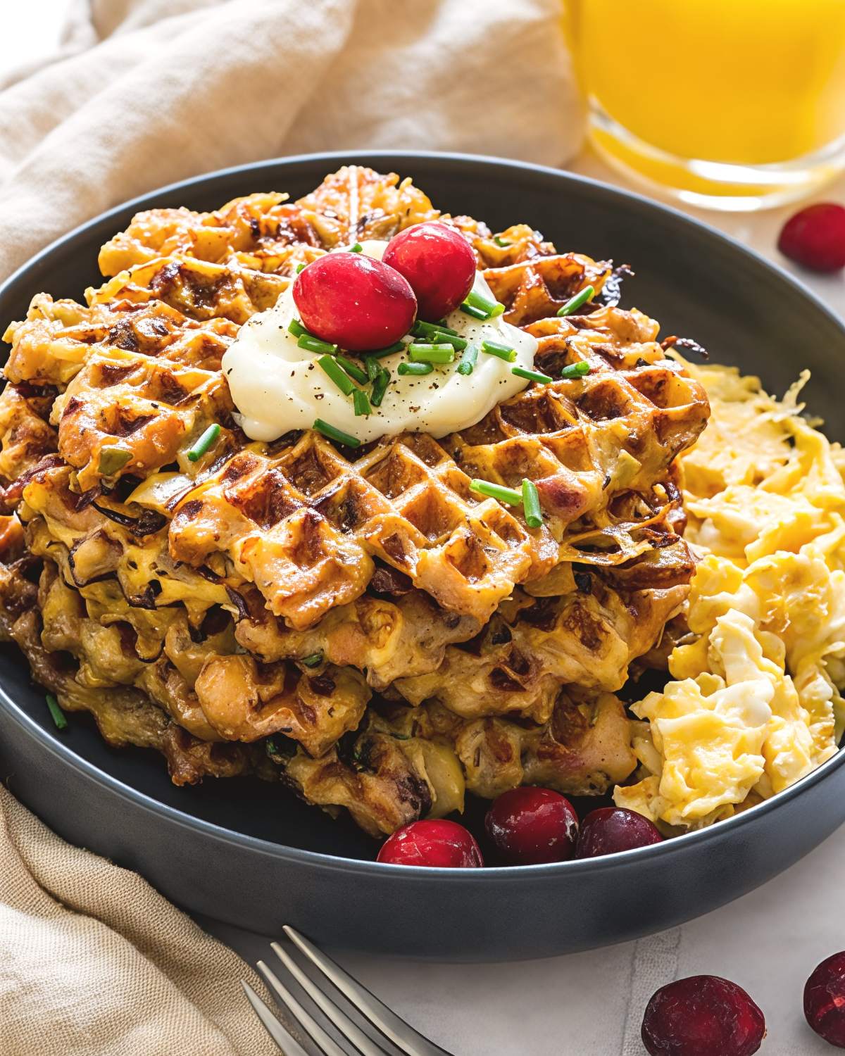 A stack of crispy Thanksgiving leftover stuffing waffles topped with mashed potatoes, fresh chives, and cranberries, served alongside scrambled eggs and orange juice.