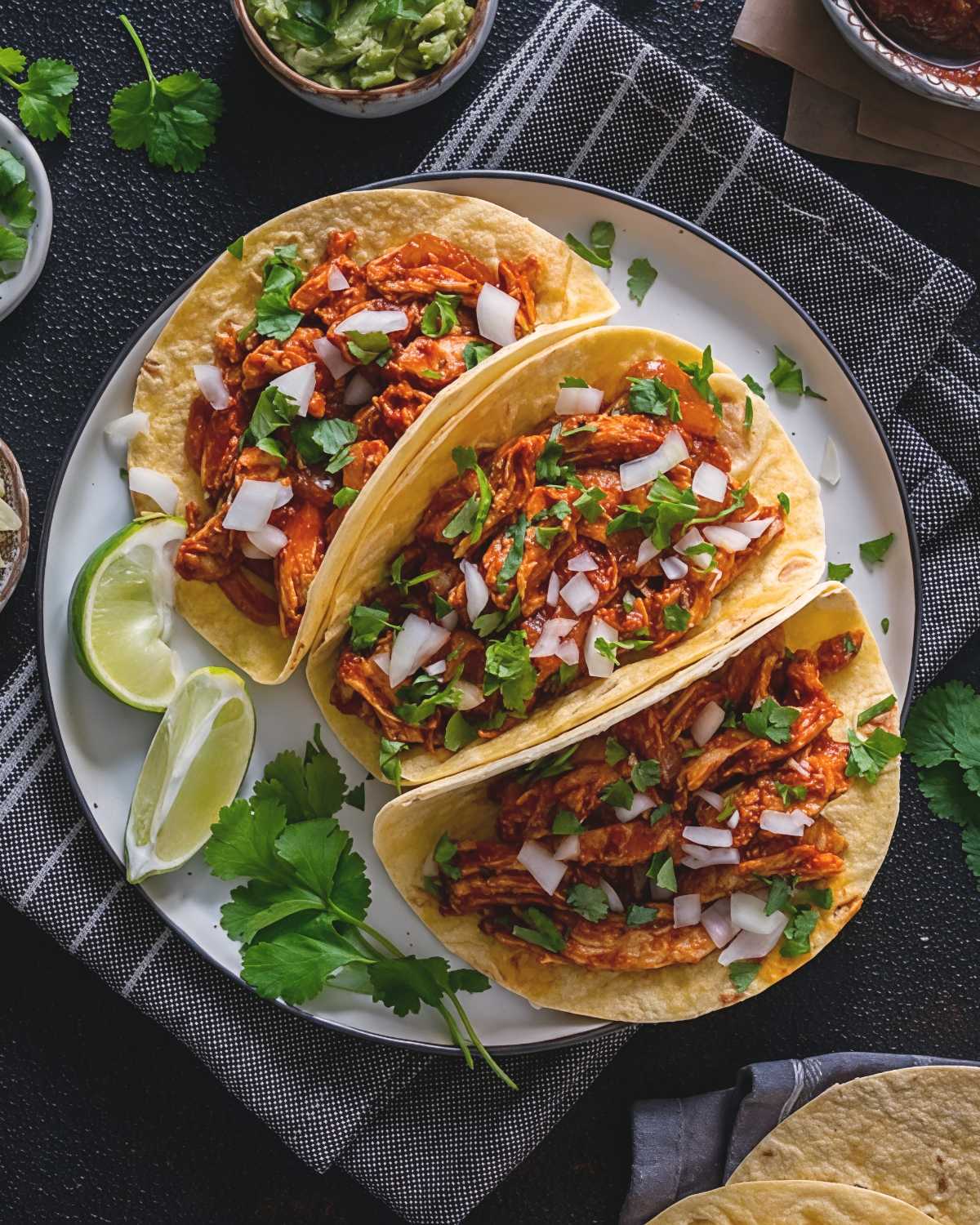 Three soft corn tortillas filled with shredded Thanksgiving leftover turkey in red sauce, topped with chopped onions and fresh cilantro on a white plate with lime wedges